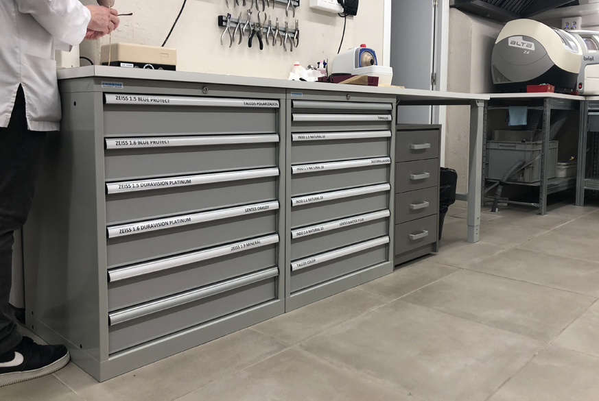 Diseño, transporte y montaje de un banco de trabajo modular compuesto por cajoneras metálicas inferiores con cajones de extracción telescópica y encimera de madera con recubrimiento laminado ubicado en la trastienda de una óptica.